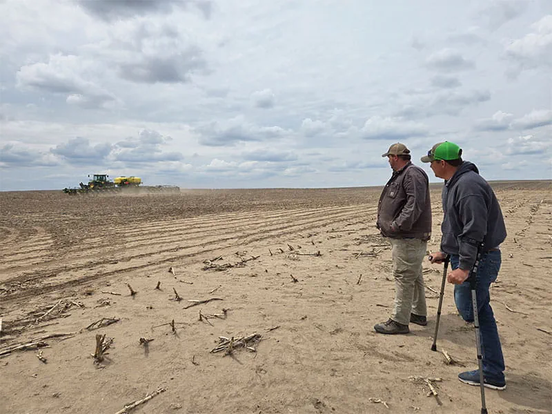 Two men, one missing his left leg, stand in a field watching a tractor and planter operate