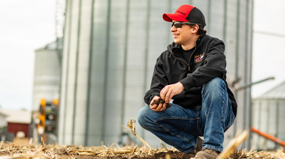 Man in hat and sunglasses in front of grain bins.