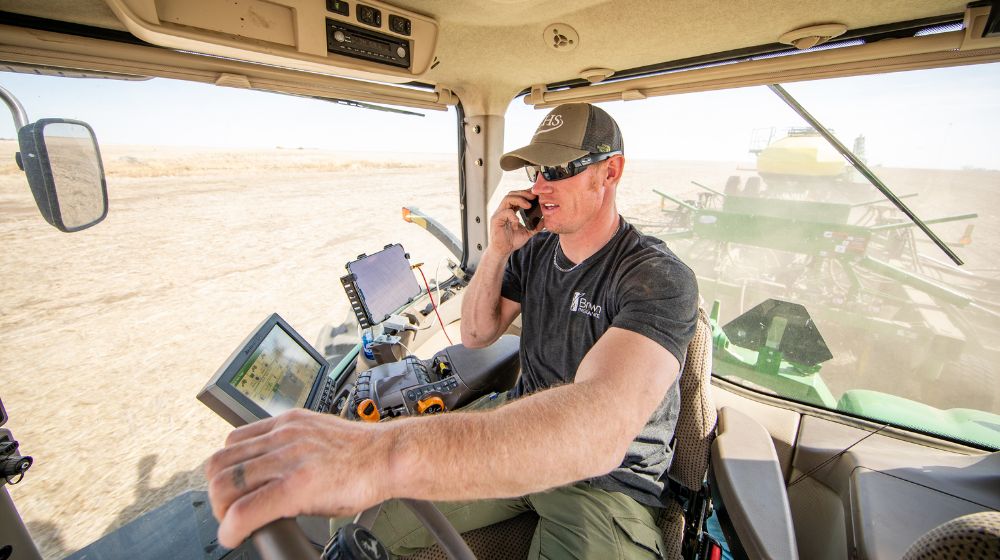 Man in hat and sunglasses in tractor cab, talking on phone.