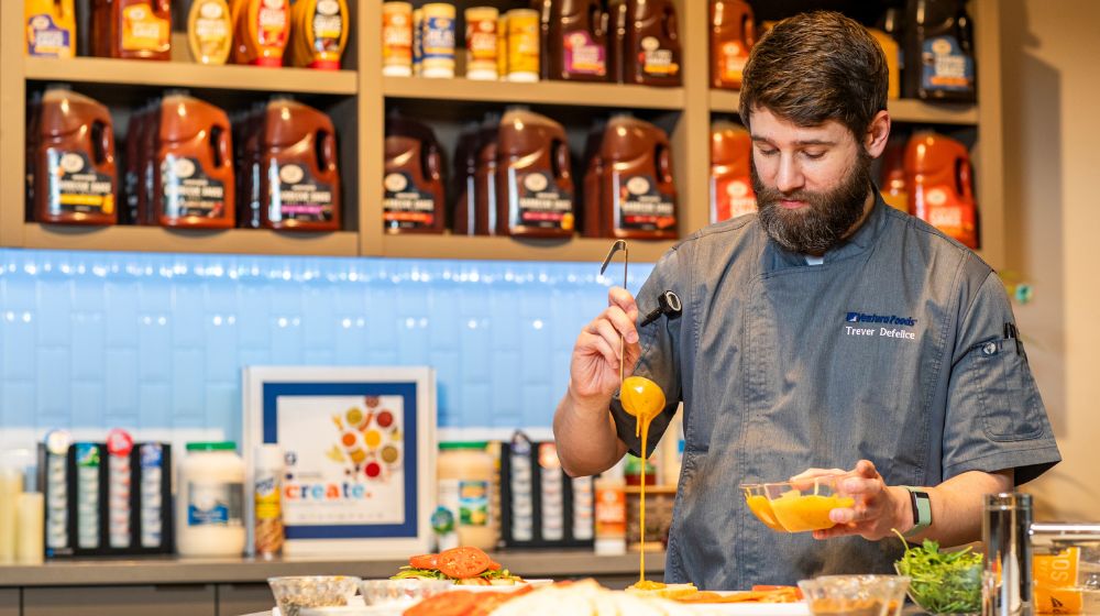 Chef drizzles sauce on a sandwich in a test kitchen lined with condiment products